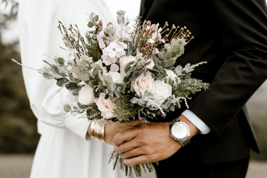Brautpaar reicht sich die Hände mit wunderschönem Brautstrauß. Heiraten in zeiten von Corona.